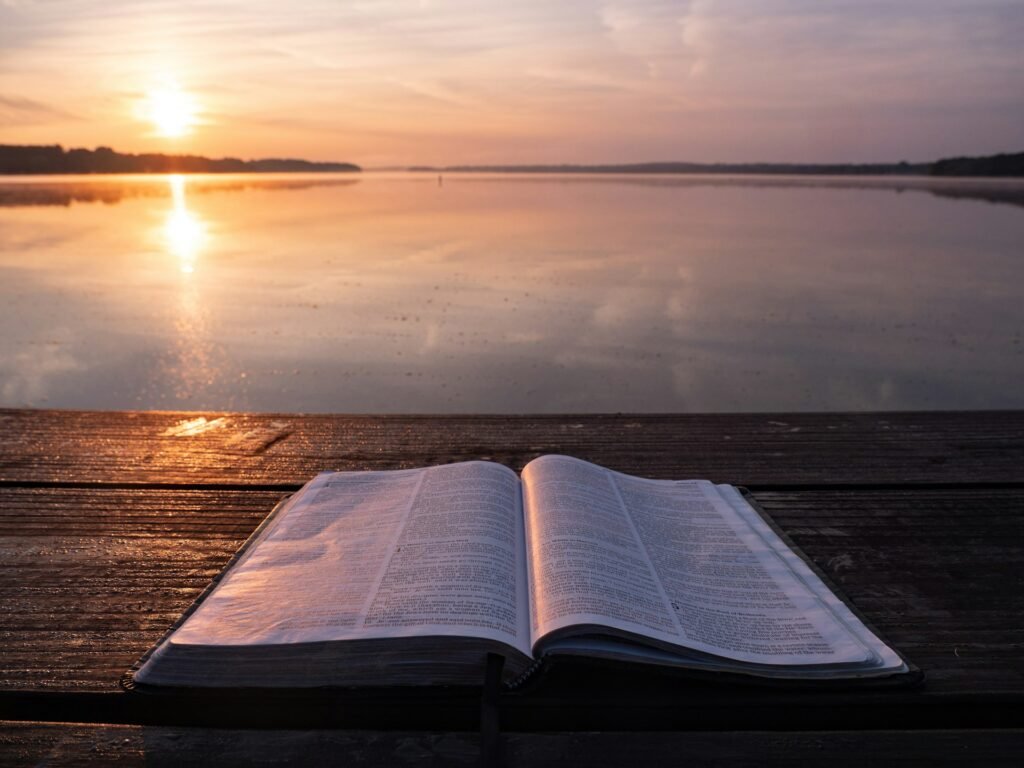An open Bible on a table beside calm water at sunrise